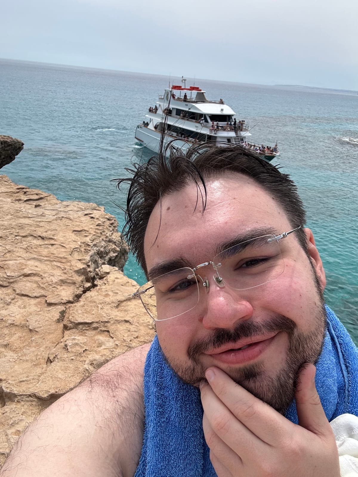 Dennis am Meer: Selfie an der Küste, ein Handtuch um den Hals, im Hintergrund ein Ausflugsboot